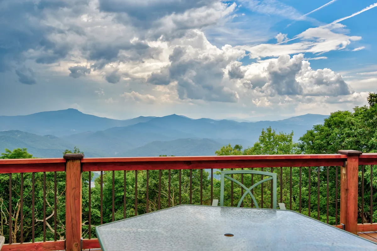A tranquil mountain landscape stretches beyond a deck railing, with comfortable seating inviting visitors to relax and soak in the scenery. The sky is broad, ideal for both sunrise watching and stargazing at night, capturing the essence of peaceful outdoor moments.
