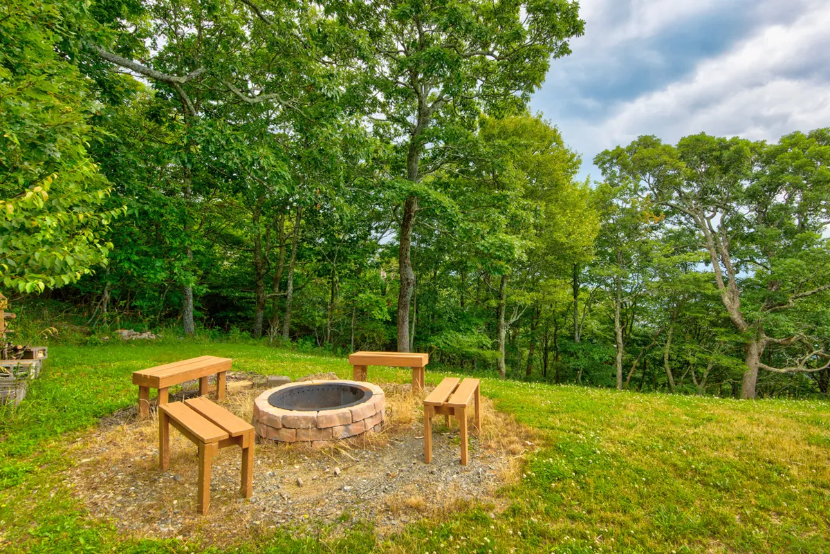 A cozy firepit surrounded by wooden benches is nestled among trees, setting the stage for intimate campfire gatherings, storytelling, and stargazing. The area feels inviting and secluded, perfect for roasting s'mores and sharing evening tales.
