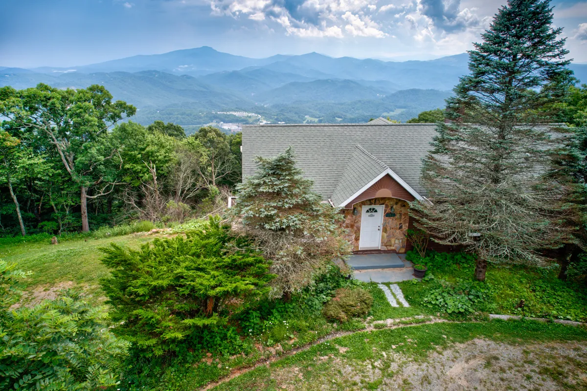 The front door and stone facade of the home are framed by mature trees and set against a serene mountain backdrop. The entryway is inviting and suggests a seamless transition from outdoor adventure to comfortable home base, tying together the idea of returning from Boone to a welcoming retreat.