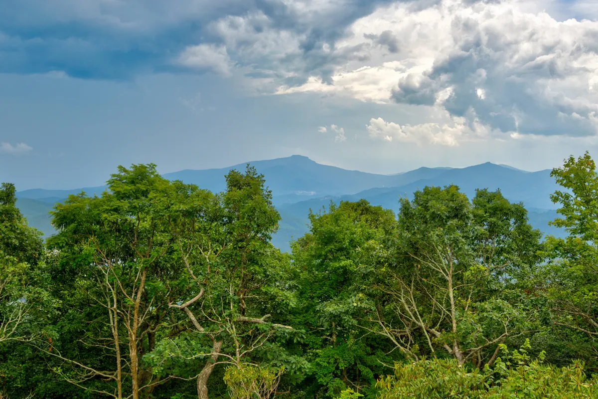 Expansive views of distant mountains and lush greenery beneath a cloudy sky set a tranquil, immersive mountain scene. The panorama highlights the area’s natural beauty and the sense of escape it provides.