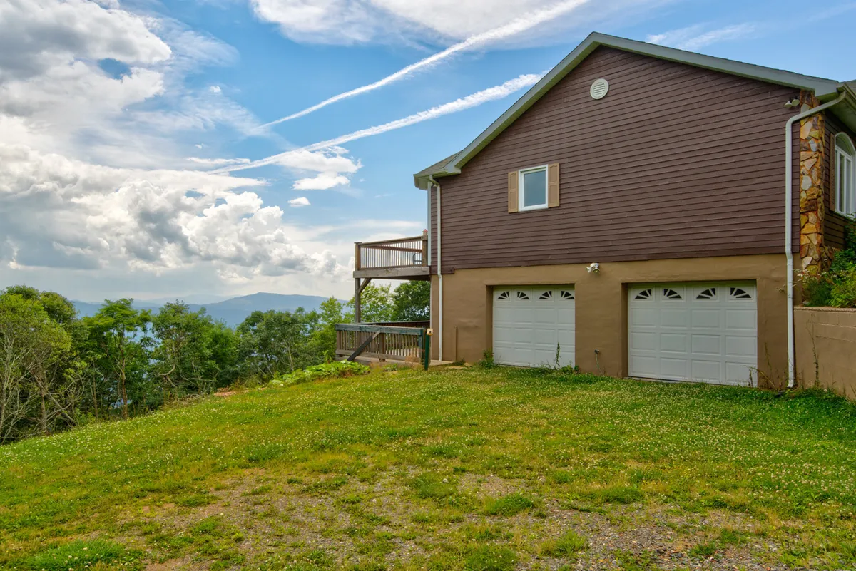 A welcoming view of the house with its large deck overlooking stunning mountain vistas. The deck is furnished for relaxation and gatherings, hinting at family activities like barbecues and enjoying sunsets. The setting is inviting, nestling the home in greenery while keeping the mountains in sight.