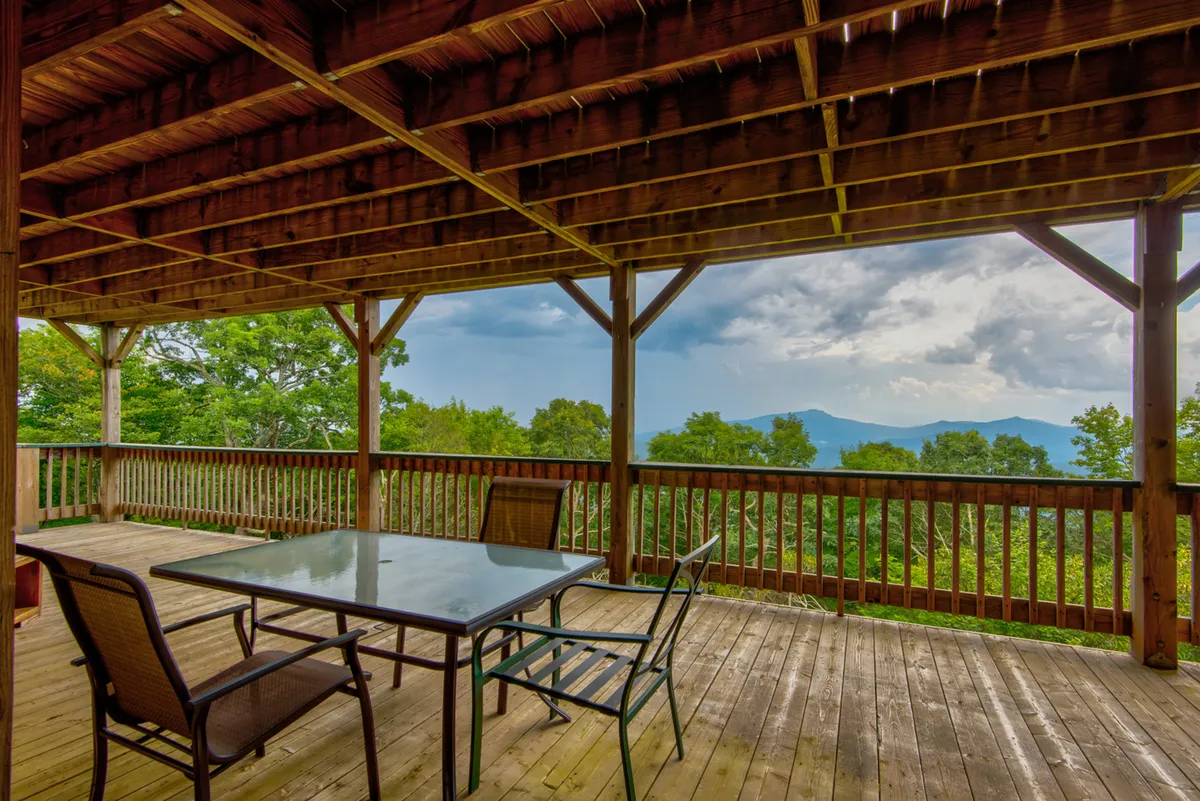 A spacious wooden deck with a dining table overlooks panoramic mountain views framed by surrounding trees. The setting is ideal for outdoor dining, relaxing, and gathering with friends or family, especially during sunset or under starlit skies. The tranquil scene hints at both morning freshness and evening serenity.