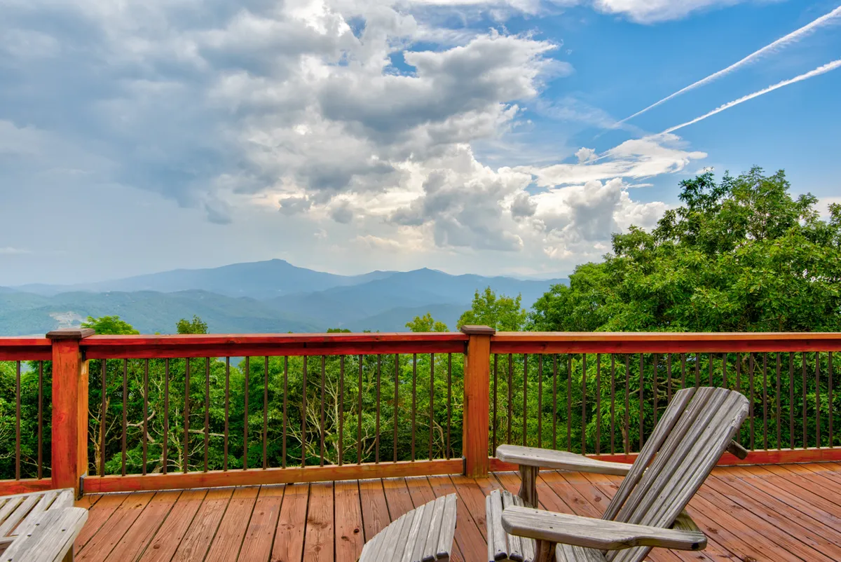 A spacious wooden deck featuring rustic railings, comfortable wooden chairs, and sweeping mountain views under a cloudy sky. The outdoor setting suggests a peaceful, contemplative spot perfect for group relaxation or evening gatherings.