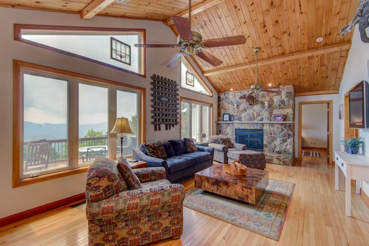 A sunlit gathering space featuring colorful furniture, a stone fireplace, and expansive windows that invite in mountain views. The wooden ceiling and coffee table complete the room's welcoming character, perfect for family gatherings and movie nights.